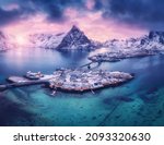 Aerial view of snowy islands with houses, rorbu, blue sea, mountains, bridge and violet cloudy sky at sunset in winter. Dramatic landscape with village, rock, road. Top view. Lofoten islands, Norway