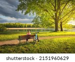 Woman sitting on bench and mountain bike, green trees and lake at sunset in spring. Colorful landscape with resting girl, bicycle, river, green grass, river in park. Summer. Sport and travel. Biking