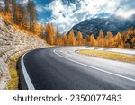 Road in mountains at sunny day in golden autumn. Dolomites, Italy. Beautiful roadway, orange tress, high rocks, blue sky with clouds. Landscape with empty highway through the mountain pass in fall