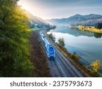 Aerial view of blue modern high speed train moving near river in alpine mountains in fog at sunrise in autumn. Top view of train, railroad, lake, reflection, trees in fall. Railway station in Slovenia