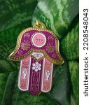 Small photo of Hamsa hand amulet (hand of Fatima or hand of Miriam) on a background of green leaves with the inscription "good luck" close-up. Decoration piece of a palm with an eye in it, used to ward off the evil