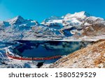 A Bernina Express train traveling along the lake shore of Lago Bianco and Piz Cambrena towering under blue sky in background after a snowfall in autumn, near Ospizio Bernina, in Grisons, Switzerland