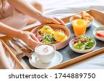 Breakfast in Bed Served with Cup of Coffee, Salad, Fresh Fruits and Eggs Benedict on Wooden Tray. Woman Hands Holding Plate with Fresh Food. Room Service in Hotel