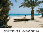 Small photo of Young woman in white shirt, shorts and sunglasses sitting on a wooden bench on a sunny sandy beach surrounded by palm trees and looking at the turquoise ocean in Santa Maria, Sal island, Cape Verde.