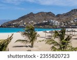 The sandy beach with palm trees, sunbeds and umbrellas, the turquoise ocean against the backdrop of mountains with houses on it and ships in harbor in Mindelo city, Sao Vicente island, Cape Verde.