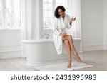 Black lady in bathrobe moisturizing smooth legs with moisturizer cream after morning shower, sitting on bathtub in light bathroom. Beauty, skincare and pampering concept