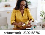 Small photo of Black woman with a chronic health condition sits at her kitchen table taking her medication. Senior woman adhering to her prescribed treatment in order to efficiently manage her personal health.