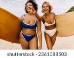 Excited female surfers going to a fun surfing spot on the beach, anticipating a happy and memorable surfing session. Two sporty women walking on shore in bikinis, eager to catch the best waves.