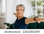 Small photo of Active senior woman doing the warrior pose at home, she stretches her arms with a confident look on her face. Mature woman practicing yoga to improve her strength, balance and stability.