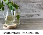 Birch sap in a transparent jug on a wooden stand. A tall glass of sap and a sprig of birch in the background. Light wooden background, space for text
