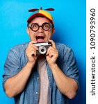 Small photo of Young nerd man with noob hat holding a vintage photo camera on blue background