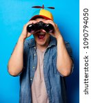 Small photo of Young nerd man with noob hat using a binoculars on blue background