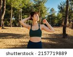 Young redhead woman touching hair and looking away, wearing green sports bra and blue yoga pants while standing on city park, outdoors. Healthy life and outdoor sports concept.
