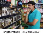 Small photo of Portrait of confused Indian man standing in front of the product counter purchasing in a grocery store. Confused man buying grocery for home in supermarket. He is thinking and totally perplexed.