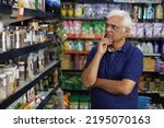 Small photo of Portrait of confused Indian old man standing in front of product counter purchasing in a grocery store. Confused old man buying grocery for home in supermarket. He is thinking and totally perplexed.