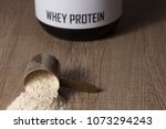 Whey protein food supplement for training and exercise. Dropped scoop with vanilla powder flavour. Wooden table. Black jar behind