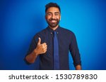 Young indian businessman wearing elegant shirt and tie standing over isolated blue background doing happy thumbs up gesture with hand. Approving expression looking at the camera showing success.