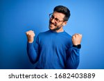 Young handsome man with beard wearing casual sweater and glasses over blue background very happy and excited doing winner gesture with arms raised, smiling and screaming for success. Celebration