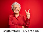 Senior beautiful grey-haired woman wearing casual shirt and glasses over red background smiling with happy face winking at the camera doing victory sign. Number two.