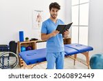 Small photo of Young hispanic man wearing physio therapist uniform reading book at clinic