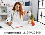 Young redhead woman nutritionist doctor at the clinic pointing down with fingers showing advertisement, surprised face and open mouth 