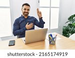Small photo of Young hispanic man with beard working at the office with laptop smiling swearing with hand on chest and fingers up, making a loyalty promise oath