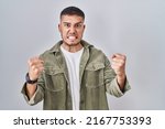 Small photo of Young hispanic man standing over isolated background angry and mad raising fists frustrated and furious while shouting with anger. rage and aggressive concept.