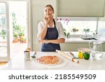 Beautiful blonde woman wearing apron cooking pizza serious face thinking about question with hand on chin, thoughtful about confusing idea 