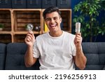 Small photo of Hispanic man holding led lightbulb and incandescent bulb smiling and laughing hard out loud because funny crazy joke.