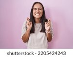 Small photo of Young hispanic woman standing over pink background gesturing finger crossed smiling with hope and eyes closed. luck and superstitious concept.