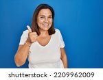 Small photo of Hispanic mature woman standing over blue background doing happy thumbs up gesture with hand. approving expression looking at the camera showing success.