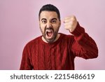 Small photo of Young hispanic man with beard wearing casual sweater over pink background angry and mad raising fist frustrated and furious while shouting with anger. rage and aggressive concept.