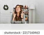 Small photo of Young latin woman sitting on the table by christmas decor gesturing finger crossed smiling with hope and eyes closed. luck and superstitious concept.