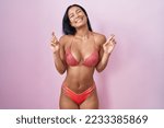 Small photo of Hispanic woman wearing bikini gesturing finger crossed smiling with hope and eyes closed. luck and superstitious concept.
