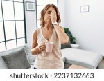 Middle age woman drinking a cup coffee at home yawning tired covering half face, eye and mouth with hand. face hurts in pain. 