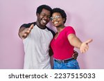 Young african american couple standing over pink background looking at the camera smiling with open arms for hug. cheerful expression embracing happiness. 
