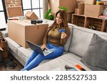 Small photo of Young latin woman using laptop and credit card sitting on sofa at hew home