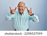 Senior man with grey hair standing over blue background shouting with crazy expression doing rock symbol with hands up. music star. heavy concept. 