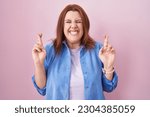 Small photo of Young hispanic woman with red hair standing over pink background gesturing finger crossed smiling with hope and eyes closed. luck and superstitious concept.