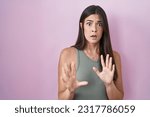 Hispanic woman standing over pink background afraid and terrified with fear expression stop gesture with hands, shouting in shock. panic concept. 