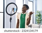 African man with dreadlocks recording vlog tutorial with smartphone at home pointing thumb up to the side smiling happy with open mouth 