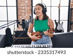 Young hispanic woman musician singing song playing ukelele at music studio