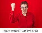Young hispanic woman with red hair standing over red background angry and mad raising fist frustrated and furious while shouting with anger. rage and aggressive concept. 