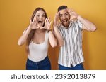 Small photo of Young couple standing over yellow background smiling cheerful playing peek a boo with hands showing face. surprised and exited