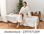 Young beautiful hispanic woman wearing bathrobe sitting on massage table at beauty salon