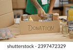 Small photo of Tattooed, hispanic man's gracious hands volunteer, putting canned food donations in a cardboard box at a bustling charity center - the beautiful side of working in altruism and unity