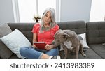 Small photo of Relaxed middle age woman, a grey-haired bibliophile, snuggled up with her loyal dog, comfortably sitting on her home sofa, indulging in the leisure of reading a riveting book in her cozy living room.