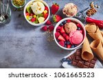 Tubs of fresh fruit with ice cream and ingredients including, chocolate, berries, walnuts, pistachio and a metal scoop for serving on slate with copy space, top down view
