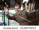 Attractive male british soldier in vintage WW2 uniform at train station, leaning out of train, waving and smiling
