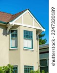 Small photo of Beige house facade with stucco exterior and white accent paint and green window decals with visible brown roof and front yard foliage. Back yard tree with clear blue sky background in neighborhood.